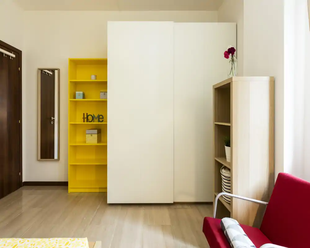Espace de vie lumineux et moderne avec une armoire blanche, une étagère jaune et un fauteuil rouge ; sol en bois propre et éléments décoratifs apportant une ambiance accueillante.