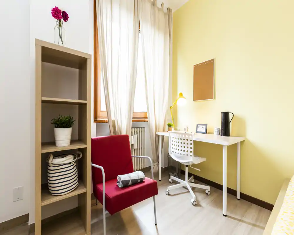 Coin de chambre compact et lumineux avec un bureau, un fauteuil rouge et des étagères. La lumière naturelle traverse les voilages et les murs jaunes, la lampe de bureau contribuant à une atmosphère chaleureuse.