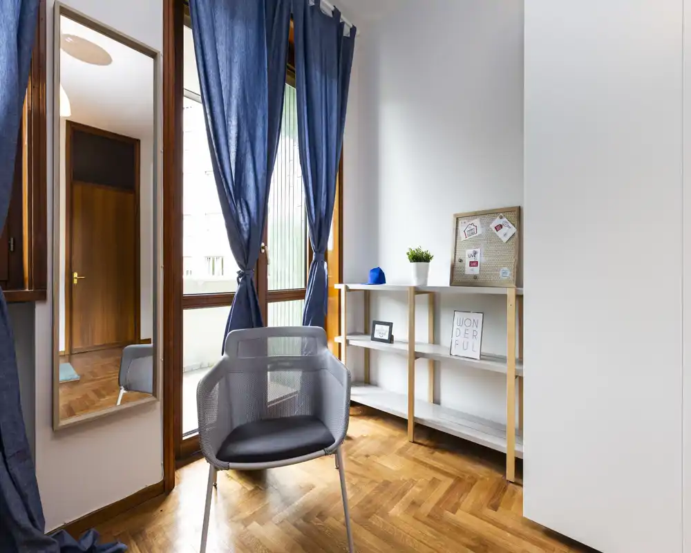 Zona giorno luminosa e minimal con poltrona in rete davanti a una grande finestra, tende blu, scaffalatura in legno e pavimento in parquet — un angolo tranquillo per leggere o rilassarsi.