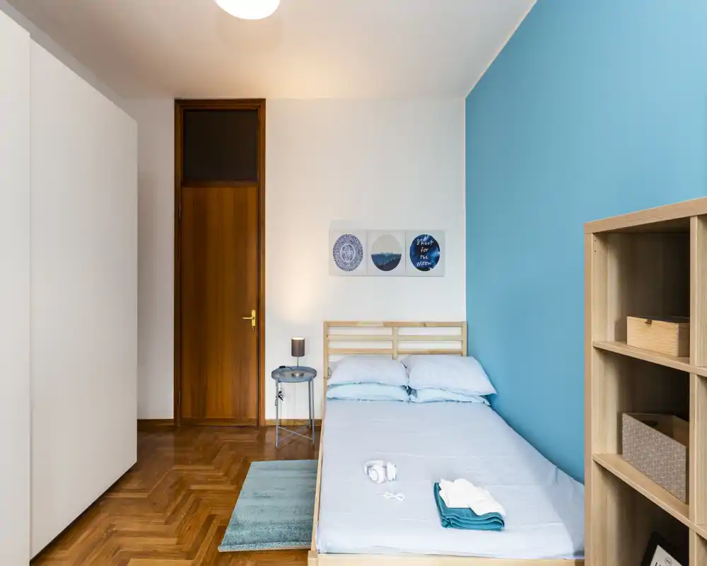 Camera da letto luminosa e ordinata con letto matrimoniale, biancheria azzurro chiaro, pavimento in legno e mensole minimaliste; stampe decorative e un comodino creano un’atmosfera moderna e tranquilla.