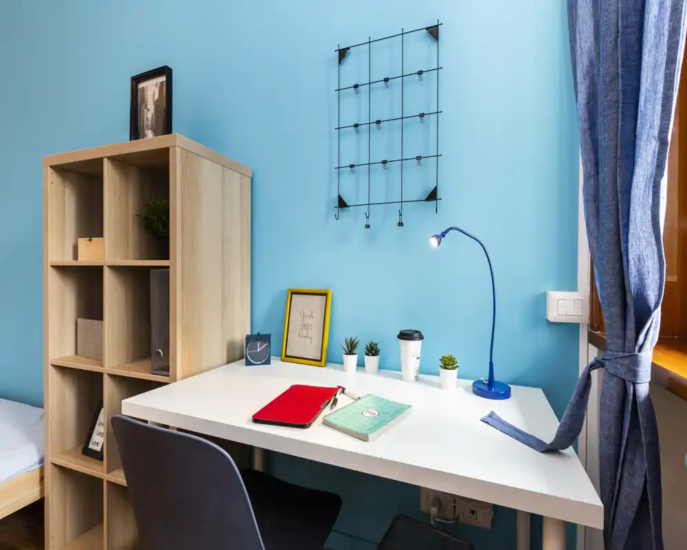Angolo camera compatto con scrivania bianca, libreria a cubi e letto parzialmente visibile sul lato sinistro; parete turchese, lampada da scrivania e piccole piante rendono l'ambiente luminoso e funzionale.
