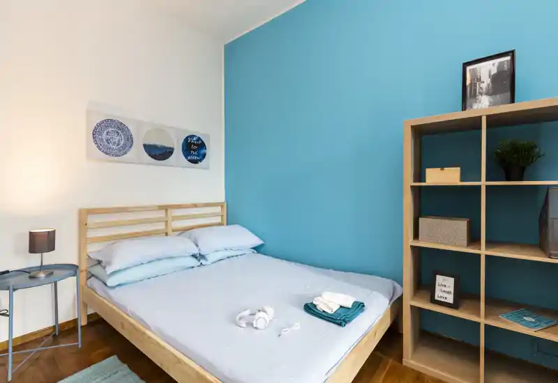 Camera da letto luminosa e ben curata con letto matrimoniale in legno, biancheria chiara e una libreria davanti a una parete turchese.