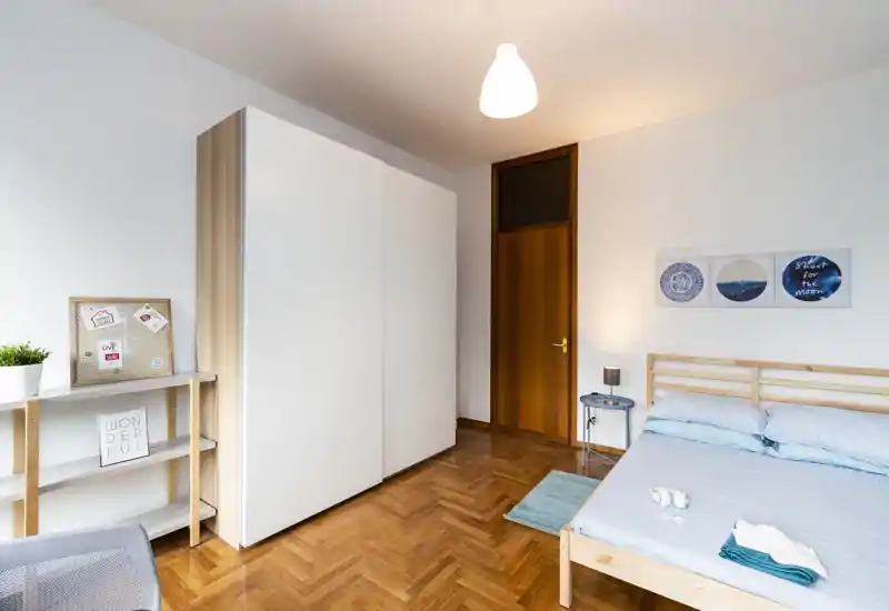 Camera da letto luminosa e ordinata con struttura del letto in legno, lenzuola azzurre, grande armadio bianco e pavimento in parquet caldo — perfetta per annunci che valorizzano comfort e spazio di archiviazione.