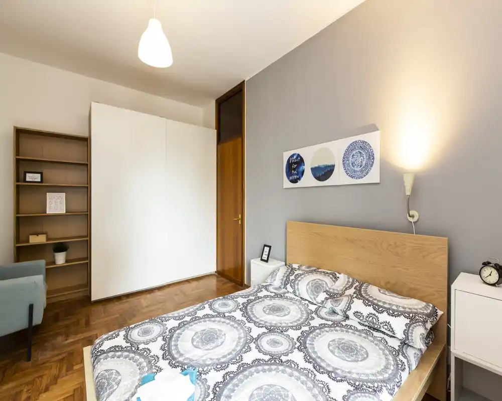 Dormitorio moderno y luminoso con cama doble, ropa de cama estampada, mesillas y lámpara acogedora; suelo de madera y un amplio armario para almacenamiento práctico.
