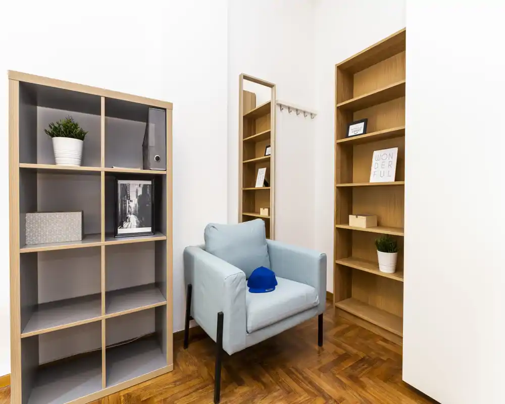 Rincón luminoso y minimalista con un cómodo sillón azul claro, estanterías abiertas, un espejo y plantas decorativas — perfecto como zona de lectura tranquila.