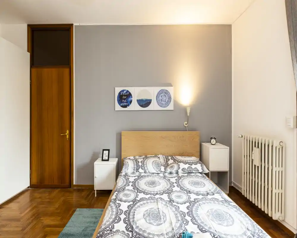 Dormitorio luminoso y bien presentado con cama doble, ropa de cama decorativa, mesitas y lámpara de pared cálida — ideal para anuncios.