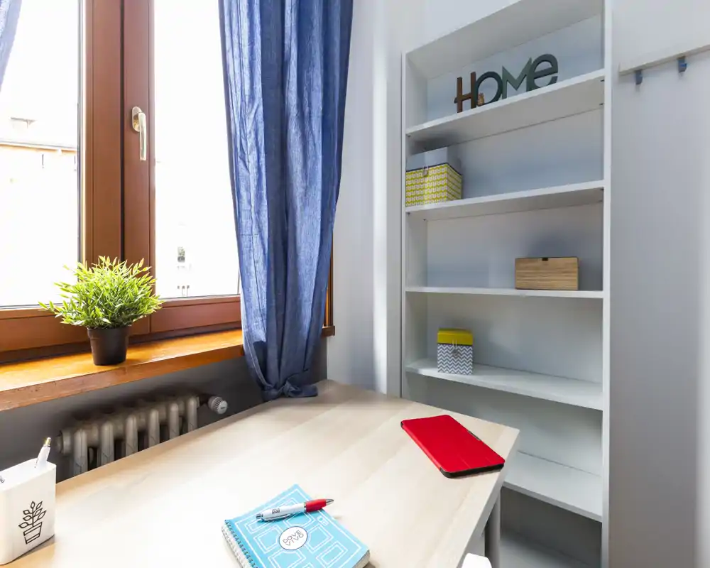 Angolo luminoso e minimale per lavoro/soggiorno con scrivania in legno vicino a una grande finestra, tenda blu e scaffalatura per riporre oggetti — perfetto per smart working o studio.