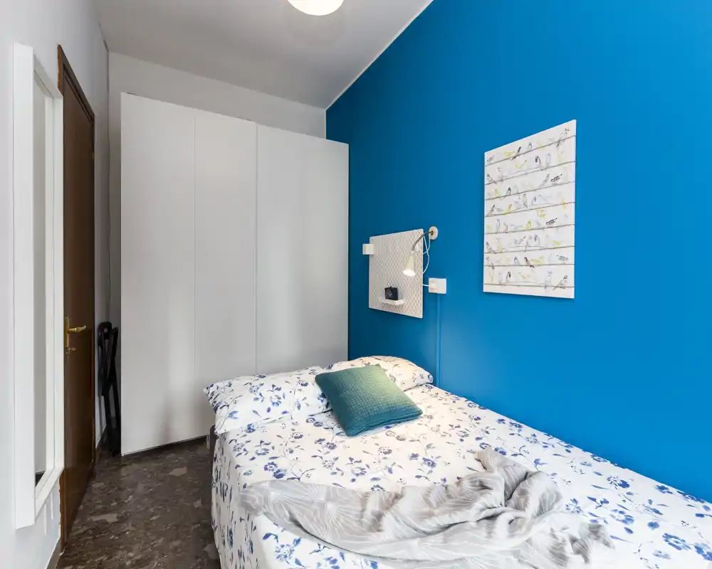 Camera compatta e ben arredata con parete azzurra a contrasto, letto matrimoniale con biancheria floreale e cuscino decorativo; ampia armadiatura bianca e luce da comodino rendono l’ambiente luminoso e accogliente.