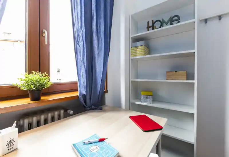 Angolo luminoso e minimale per lavoro/soggiorno con scrivania in legno vicino a una grande finestra, tenda blu e scaffalatura per riporre oggetti — perfetto per smart working o studio.