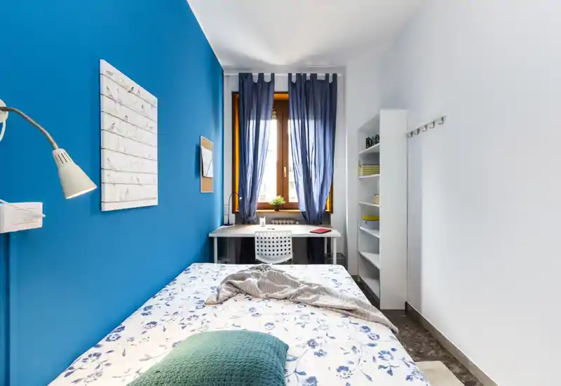 Camera da letto luminosa e compatta con parete blu, letto singolo con lenzuola floreali e una scrivania vicino alla finestra — ideale per studenti o soggiorni brevi.