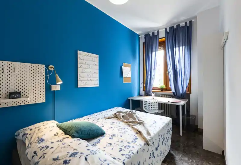 Camera singola luminosa e stilosa con copriletto a fiori, cuscino turchese e parete d'accento blu intenso. Una scrivania compatta vicino alla finestra sfrutta la luce naturale, perfetta per studiare o lavorare da remoto.