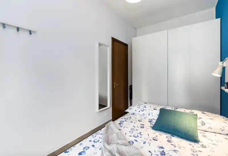 Camera da letto luminosa e compatta con copriletto floreale, cuscino verde acqua e un ampio armadio bianco. L'inquadratura valorizza l'area notte e lo spazio contenitivo, ideale per una persona o una coppia.