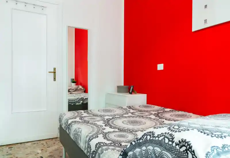 Dormitorio compacto y luminoso con cama doble estampada y una pared roja de acento; incluye mesita de noche y espejo de cuerpo entero que refleja la cama.