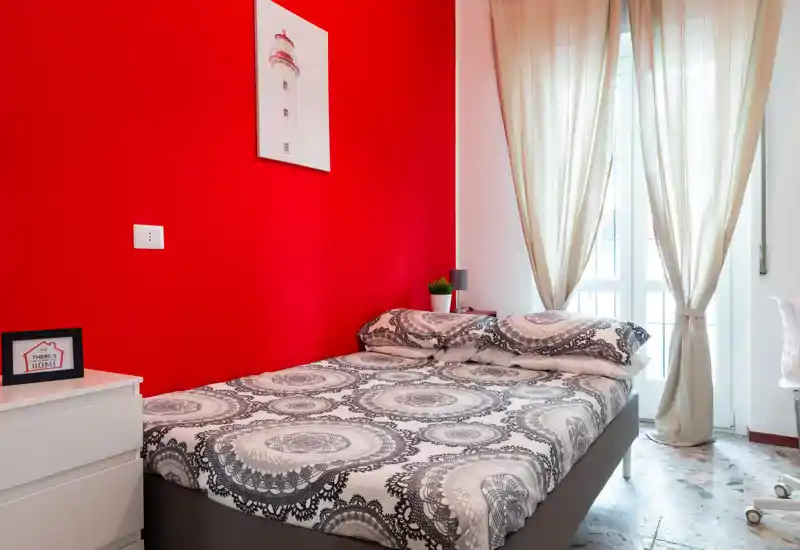 Dormitorio moderno y luminoso con cama doble y ropa de cama estampada, pared de acento roja y cortinas claras que dejan pasar la luz natural. Habitación limpia y acogedora con lámpara de noche y planta.
