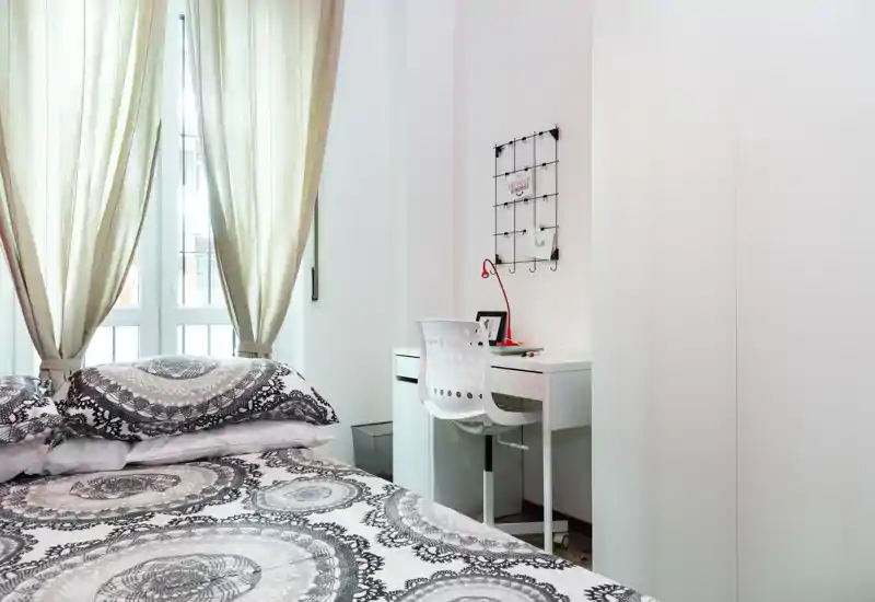 Dormitorio luminoso y bien decorado con ropa de cama estampada, luz natural suave desde la ventana y un pequeño escritorio blanco con silla—ideal para una estancia cómoda y compacta.