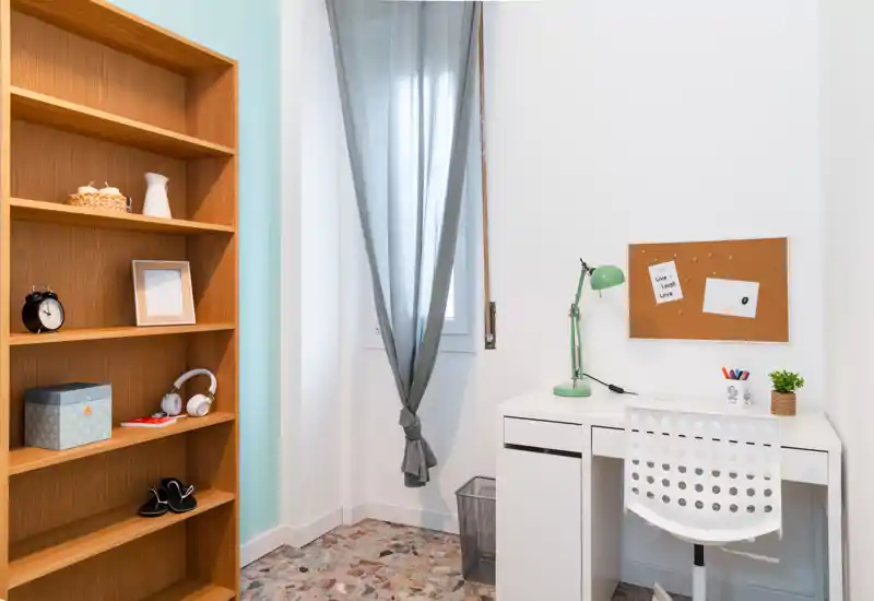 Pequeño espacio de trabajo luminoso y ordenado con escritorio blanco, silla perforada y una estantería de madera — ideal para estudiar o teletrabajar junto a una ventana con cortinas suaves.