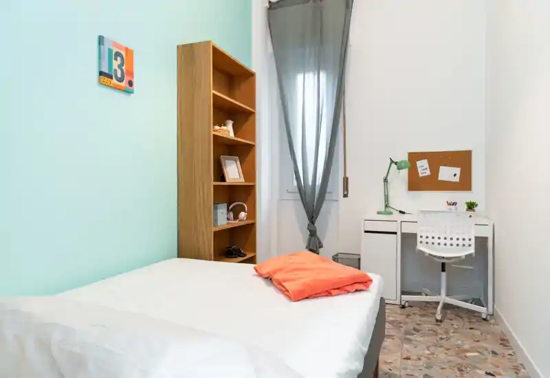 Dormitorio individual luminoso y ordenado con cama cómoda, estantería y un pequeño escritorio junto a la ventana — ideal para estudiante o una sola persona.