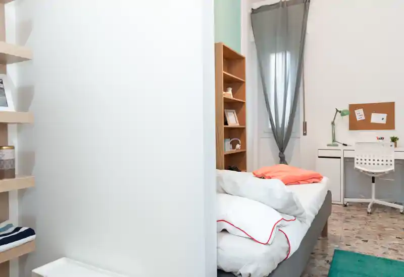 Dormitorio luminoso y ordenado con cama bien hecha, estantería y un pequeño escritorio junto a la ventana — ideal para una persona o estudiante.