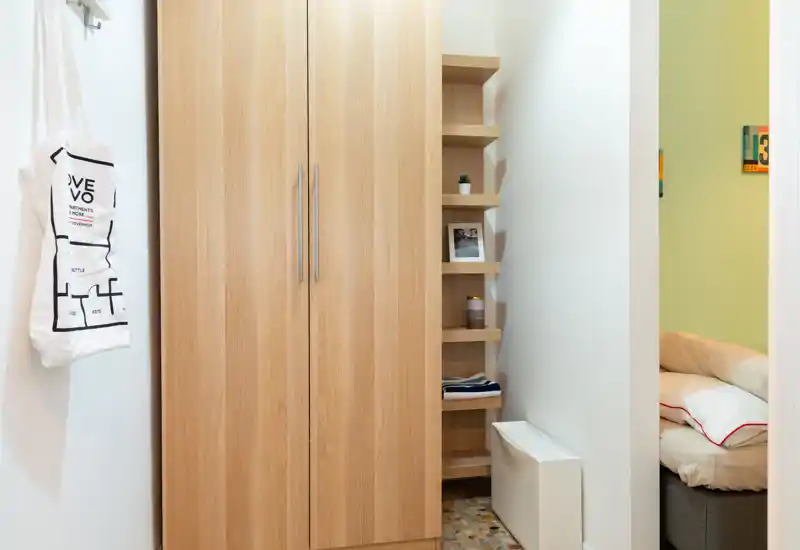 Pequeño pasillo con armario y estanterías en primer plano; a través de la puerta se ve parcialmente una cama.