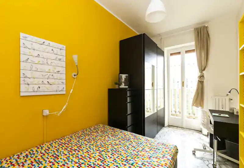 Camera da letto moderna e luminosa con parete gialla, copriletto colorato, armadio con ante a specchio e scrivania vicino alla porta del balcone — ariosa e ben illuminata.