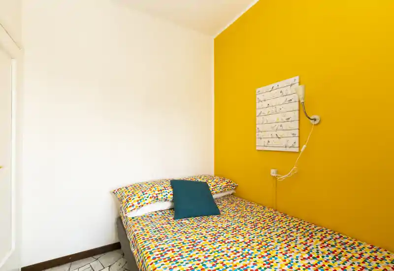 Camera da letto luminosa e colorata con letto matrimoniale contro una parete gialla vivace, biancheria a fantasia e cuscino decorativo che creano un'atmosfera accogliente e allegra.