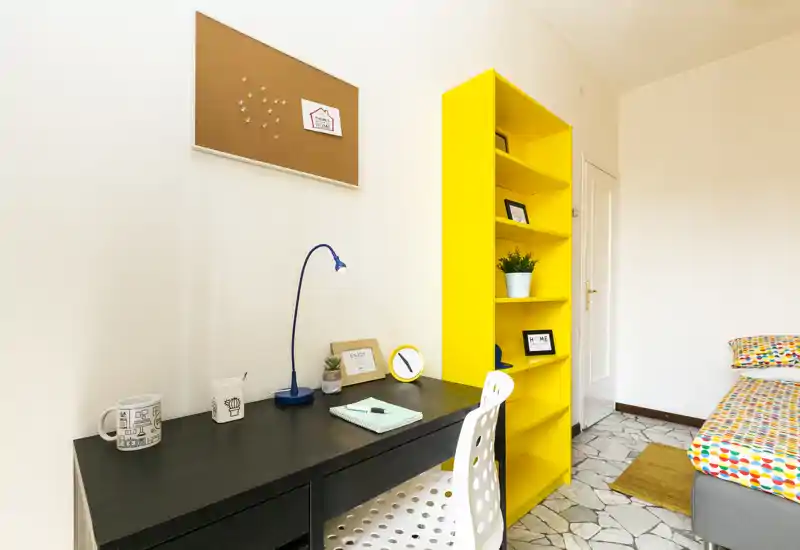 Angolo di camera luminoso e moderno con scrivania, biancheria colorata e una libreria alta gialla. L'immagine mette in evidenza un'area studio funzionale e un letto singolo accogliente, ideale per studenti o soggiorni brevi.