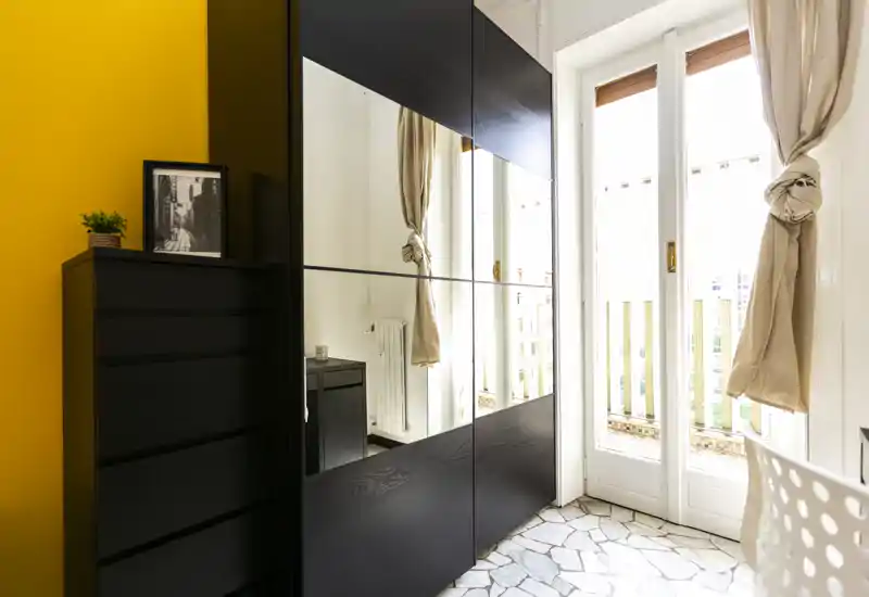 Angolo di camera luminoso con un grande armadio con ante a specchio, comò e una porta finestra che illumina l'ambiente con luce naturale.