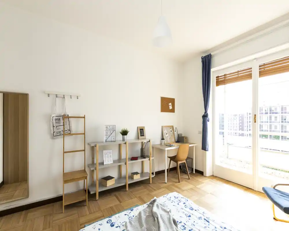 Camera da letto luminosa e minimal con letto parzialmente visibile in primo piano, pavimento in parquet, una scrivania vicino alla porta del balcone e scaffali con decorazioni curate.