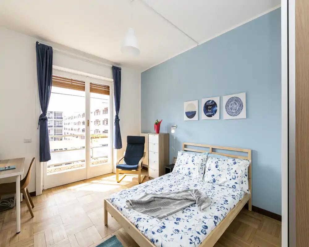Camera da letto luminosa e moderna con letto matrimoniale con biancheria floreale blu, struttura in legno chiaro, una poltrona comoda e accesso a un balcone soleggiato tramite ampie porte in vetro.