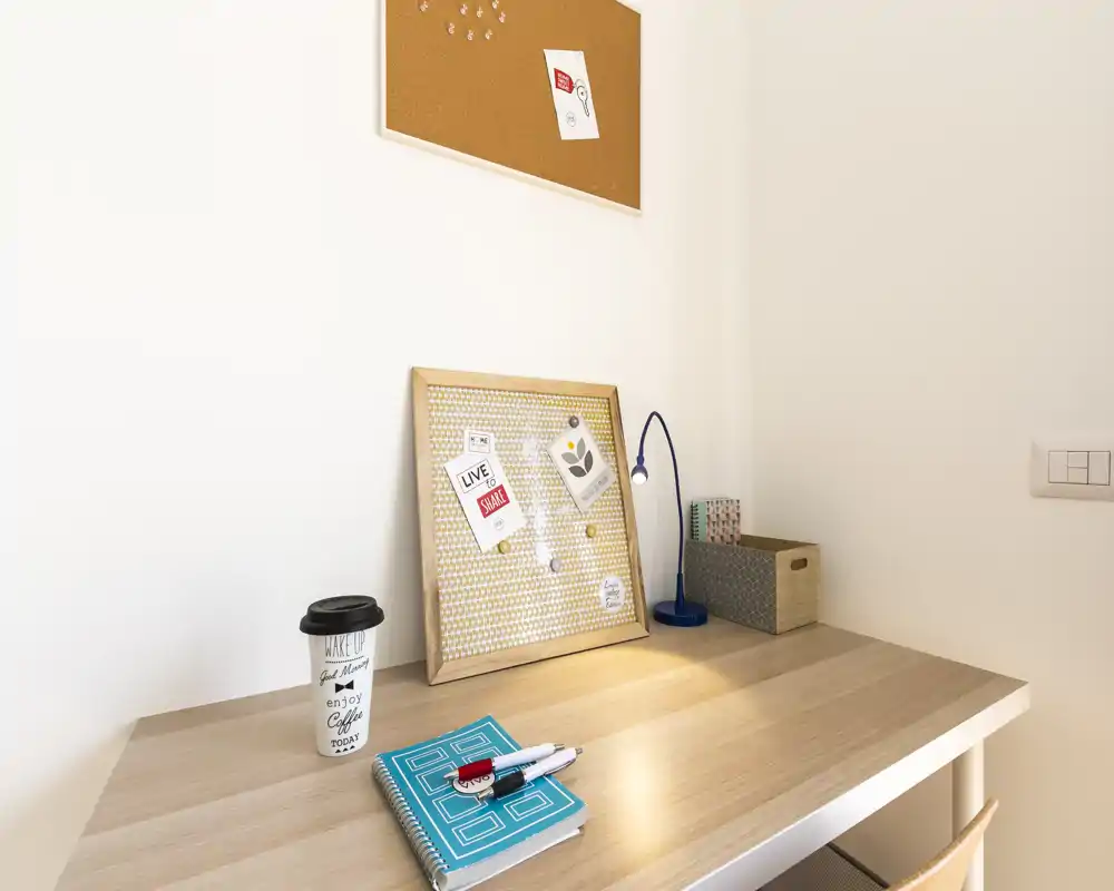 Angolo scrivania luminoso e minimal con bacheca in sughero, lampada, taccuino e bicchiere da caffè da asporto — un piccolo spazio di lavoro domestico ordinato.