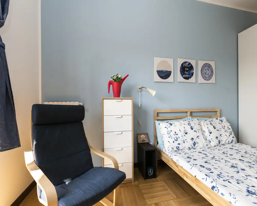 Camera da letto luminosa con struttura del letto in legno, biancheria floreale e semplici quadretti; presente una poltrona comoda e un cassettiera alta con pianta decorativa.