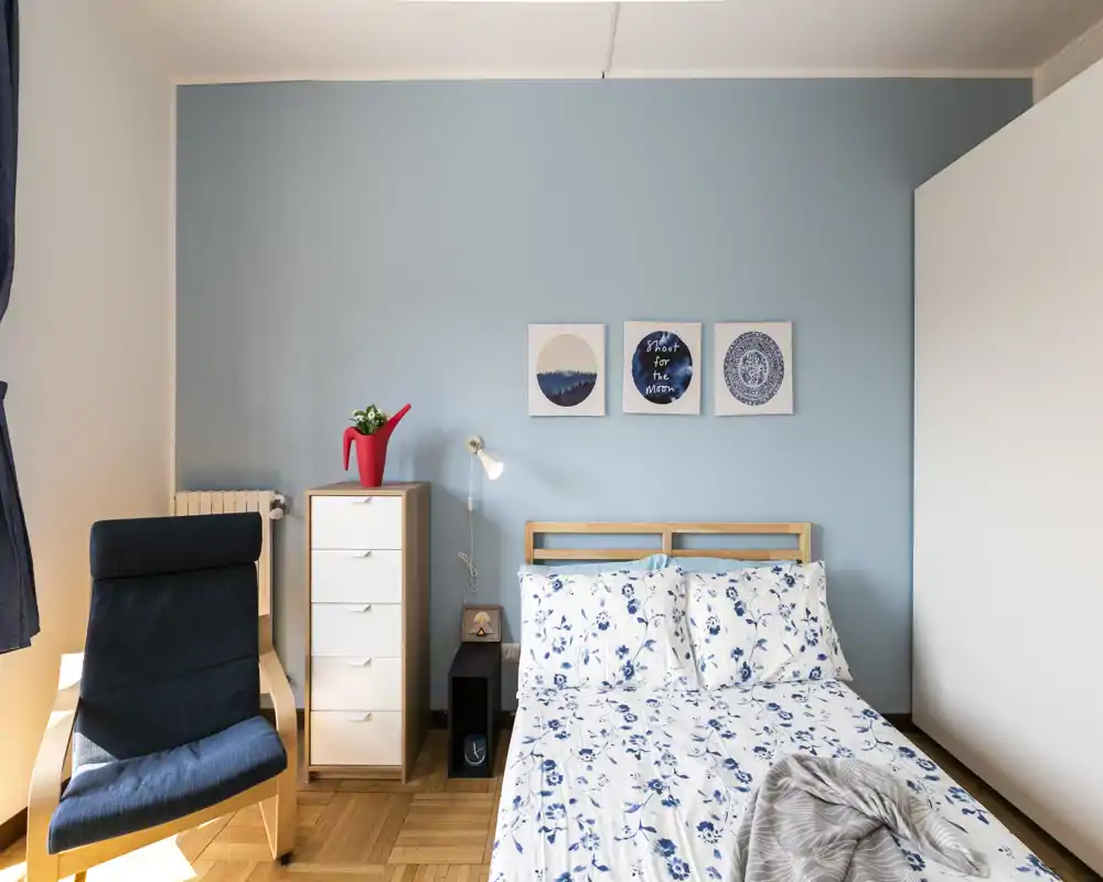 Camera da letto luminosa e ordinata con letto matrimoniale in lenzuola blu e bianche a fiori, comodino e quadri coordinati — moderna e accogliente.