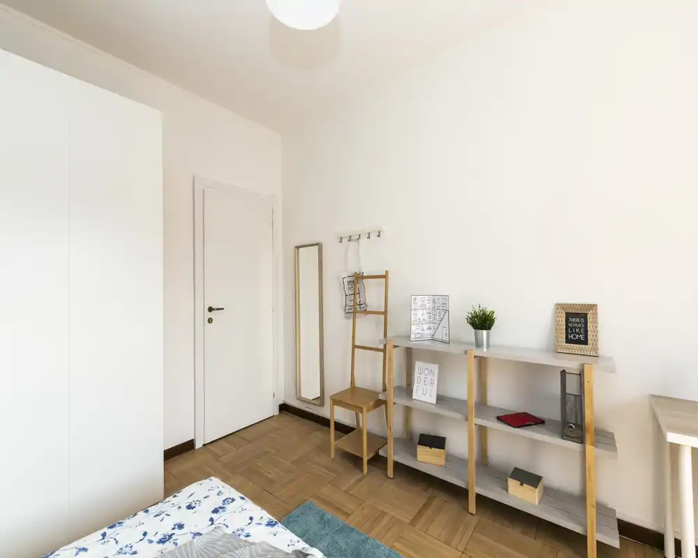 Angolo di camera da letto luminoso e compatto con la parte di un letto in primo piano, scaffalatura in legno, specchio a tutta altezza e pareti chiare per un aspetto minimal e pulito.