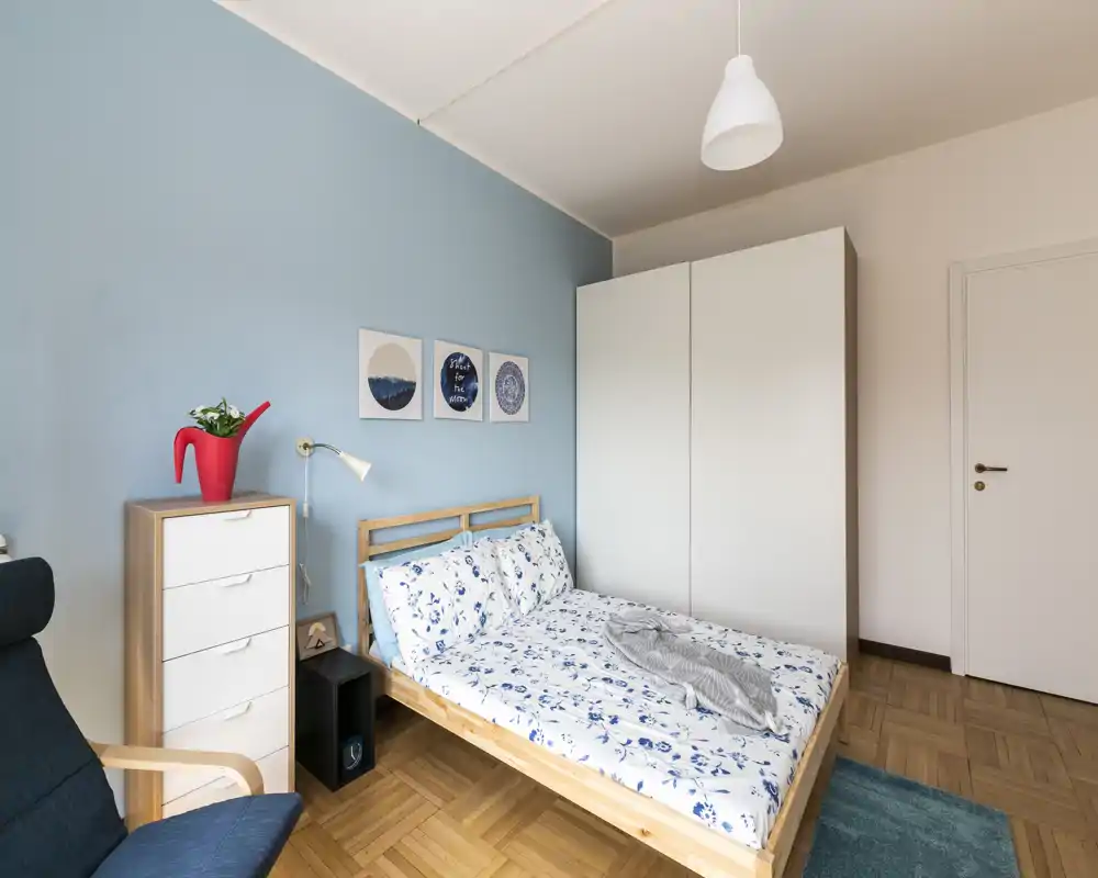 Camera da letto luminosa e ordinata con letto matrimoniale, parete blu e armadio bianco — biancheria accogliente e decorazioni essenziali per un'atmosfera rilassante.