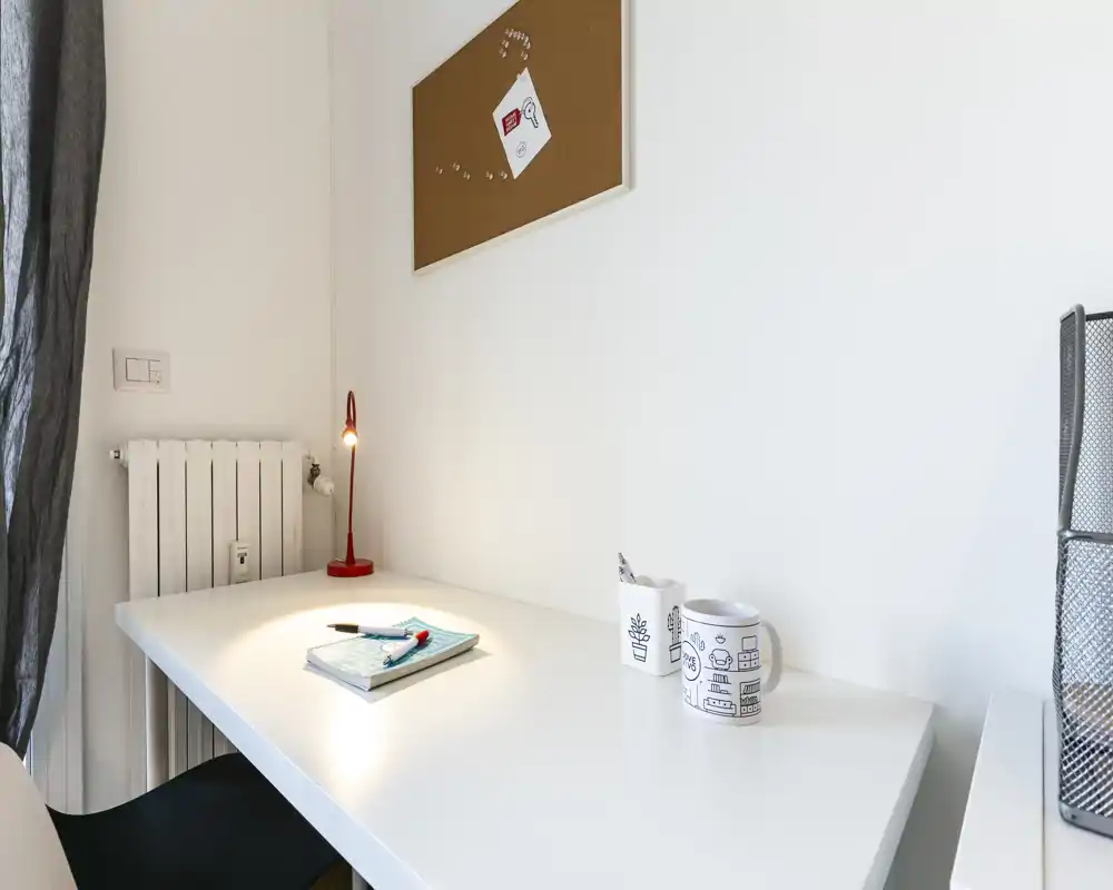 Area di lavoro luminosa e minimal in un soggiorno, con scrivania bianca, piccola lampada rossa e decorazioni essenziali — ordinata e ben illuminata.