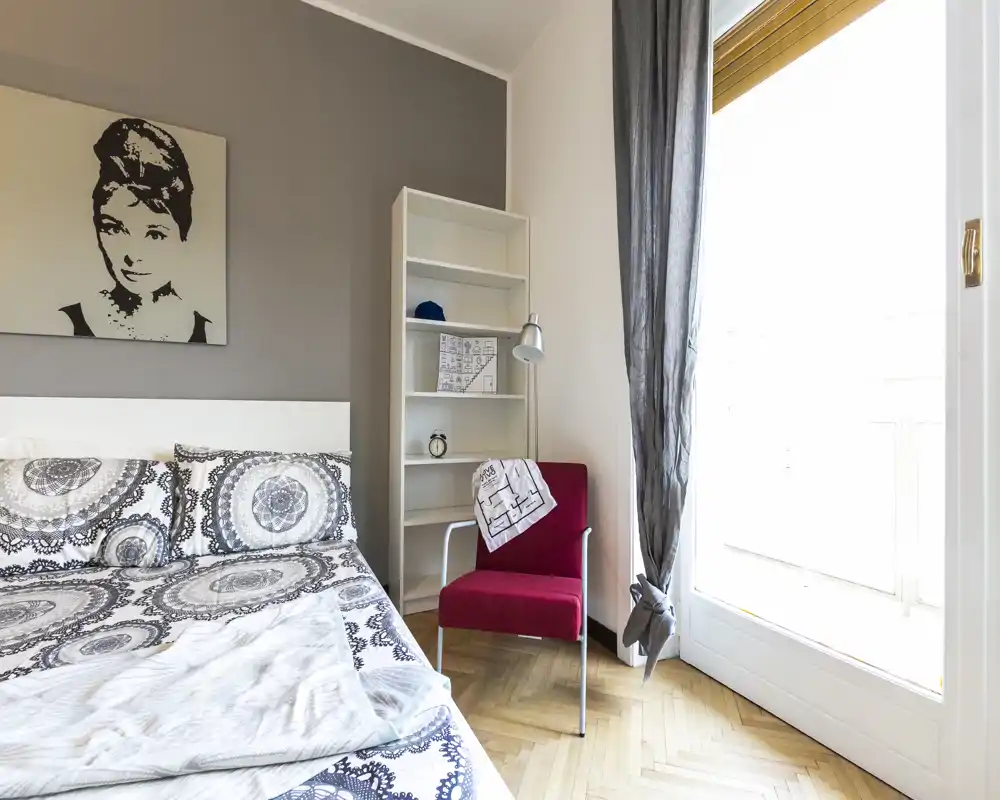 Camera da letto luminosa e curata, con biancheria a motivo, un quadro decorativo, una poltroncina rossa come elemento d'accento e una grande finestra che assicurano molta luce naturale.