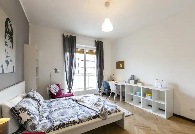 Camera da letto luminosa e moderna con letto matrimoniale, biancheria decorata, poltrona per la lettura e scrivania vicino alla finestra — ideale per un soggiorno confortevole con luce naturale.