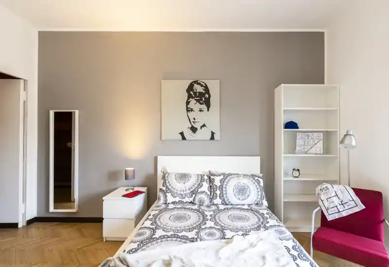 Camera da letto luminosa e di stile con letto matrimoniale centrale, biancheria coordinata, comodino con lampada, libreria e una comoda poltrona — perfetta per un annuncio moderno.