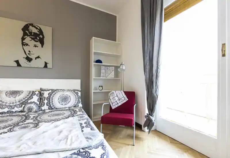 Camera da letto luminosa e curata, con biancheria a motivo, un quadro decorativo, una poltroncina rossa come elemento d'accento e una grande finestra che assicurano molta luce naturale.