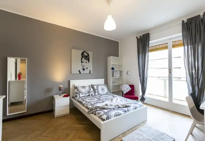 Camera da letto moderna e luminosa con letto matrimoniale, biancheria decorata, comodino con lampada, libreria e ampia finestra con tende leggere che lasciano entrare luce naturale.