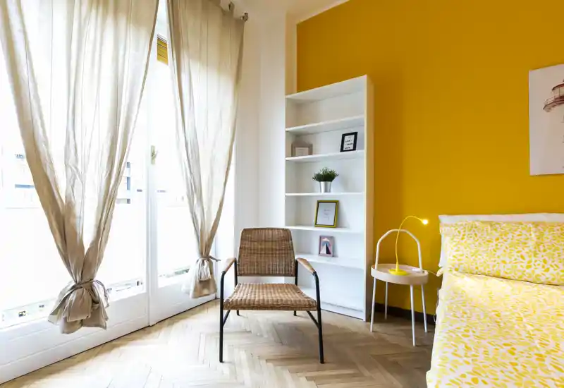 Camera da letto luminosa e moderna con piumone giallo a motivo, una poltrona in rattan e grandi finestre che lasciano entrare molta luce naturale — accogliente e di stile.