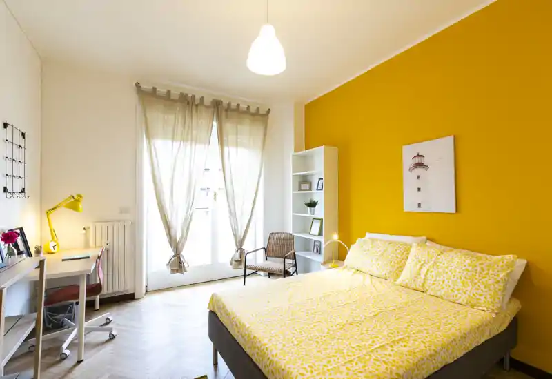 Camera da letto luminosa e di stile con parete d'accento gialla, letto matrimoniale con biancheria a fantasia, scrivania e porte sul balcone che garantiscono molta luce naturale.