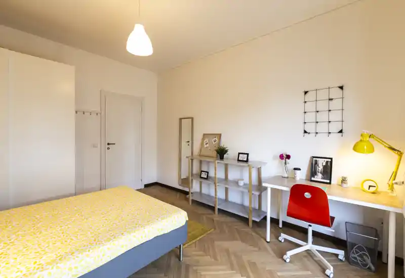 Camera da letto luminosa e ben arredata con letto matrimoniale, pavimento in parquet e un angolo studio ordinato con una lampada gialla vivace — ideale per riposo e lavoro.