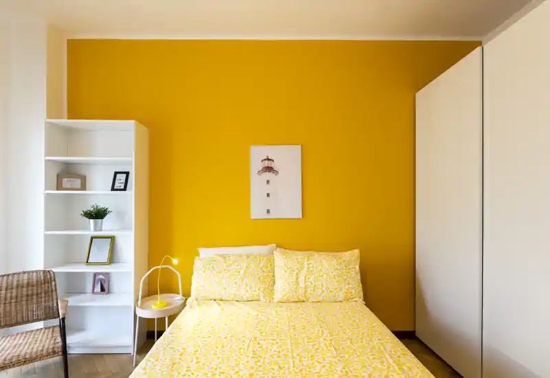 Camera da letto luminosa e curata con un letto matrimoniale con biancheria gialla a motivi su una parete color senape, lampada da comodino e scaffalatura ordinata — accogliente e solare.