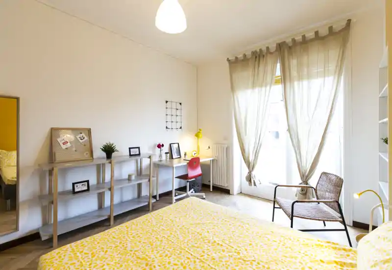 Camera da letto luminosa e ben arredata con letto matrimoniale con biancheria gialla a motivo, una scrivania con sedia per lavoro e una comoda poltrona in vimini vicino alla finestra che illumina la stanza con luce naturale.