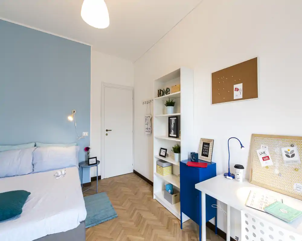 Dormitorio luminoso y moderno con cama bien hecha frente a una pared azul suave, lámpara de noche y alfombra. Una estantería y un escritorio ordenado ofrecen almacenamiento y zona de estudio, ideal para estudiante o profesional.