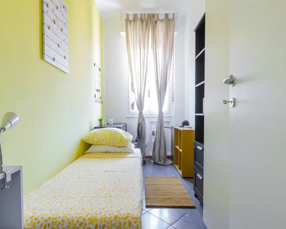 Camera singola luminosa con letto ben rifatto, dettagli gialli e luce naturale dalla finestra. Scaffali e comodino funzionali, ideale per brevi soggiorni o studenti.