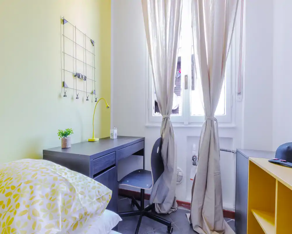 Piccola camera luminosa con letto singolo, scrivania nera e sedia vicino alla finestra. Tende neutre e accenti gialli donano un aspetto moderno e fresco — ideale per studenti o affitti brevi.