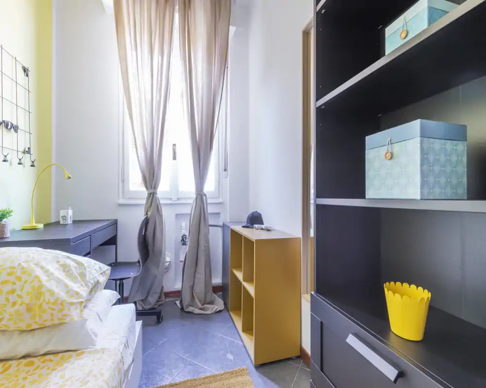Camera da letto singola, luminosa e moderna con biancheria a fantasia, una scrivania vicino alla finestra e scaffalature aperte; tende neutre lasciano entrare luce naturale.