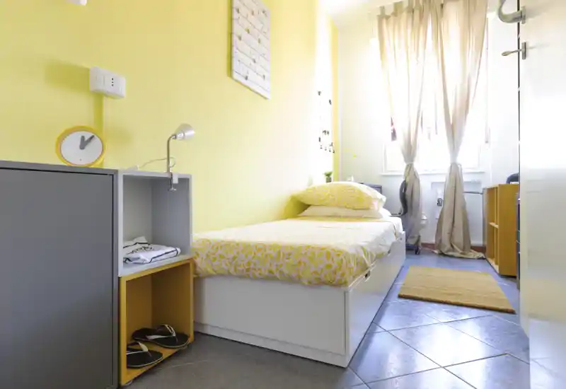 Camera singola luminosa e compatta con letto su pedana bianco, biancheria gialla a motivo e cuscini coordinati. Luce naturale intensa dalla finestra, pareti gialle calde e soluzioni contenitive abbinate rendono la stanza accogliente per un ospite singolo.